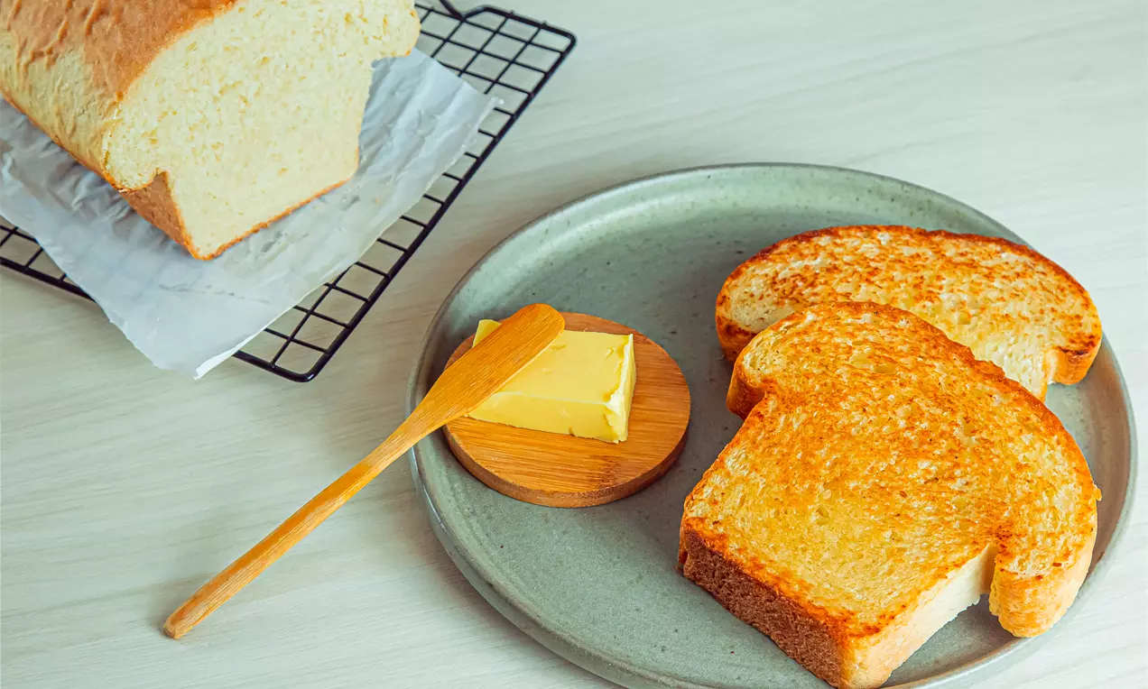 Fatias de pão caseiro douradas e macias com textura levemente tostada ao lado de uma porção de manteiga no prato cinza