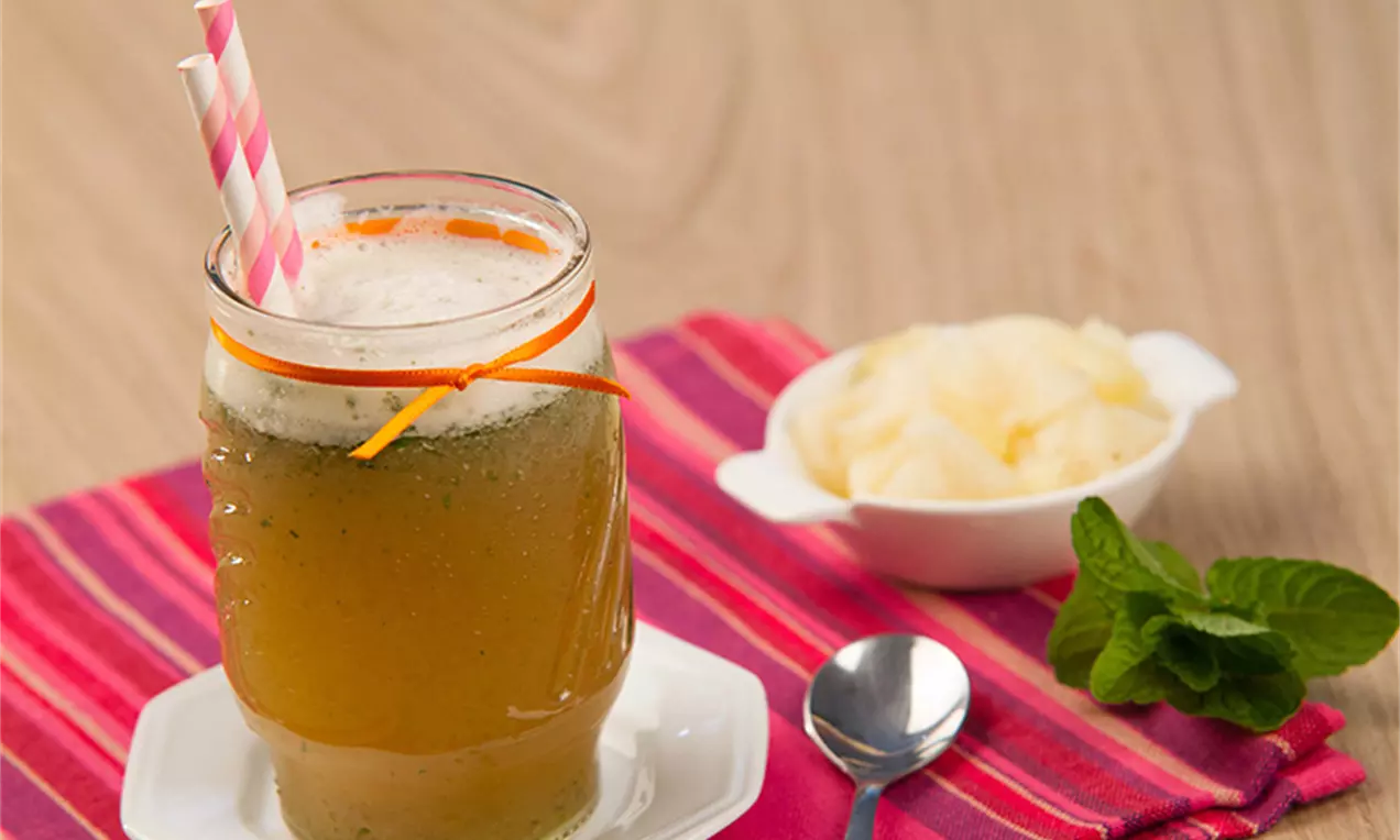 Suco dourado com espuma leve em copo de vidro decorado com raspas de laranja e canudos listrados, ao lado de hortelã, colher e abacaxi em pedaços