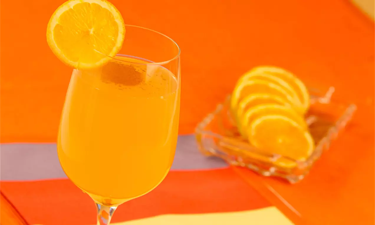 Suco laranja brilhante em uma taça fina decorada com rodela de laranja na borda, com fatias em prato quadrado de vidro ao fundo