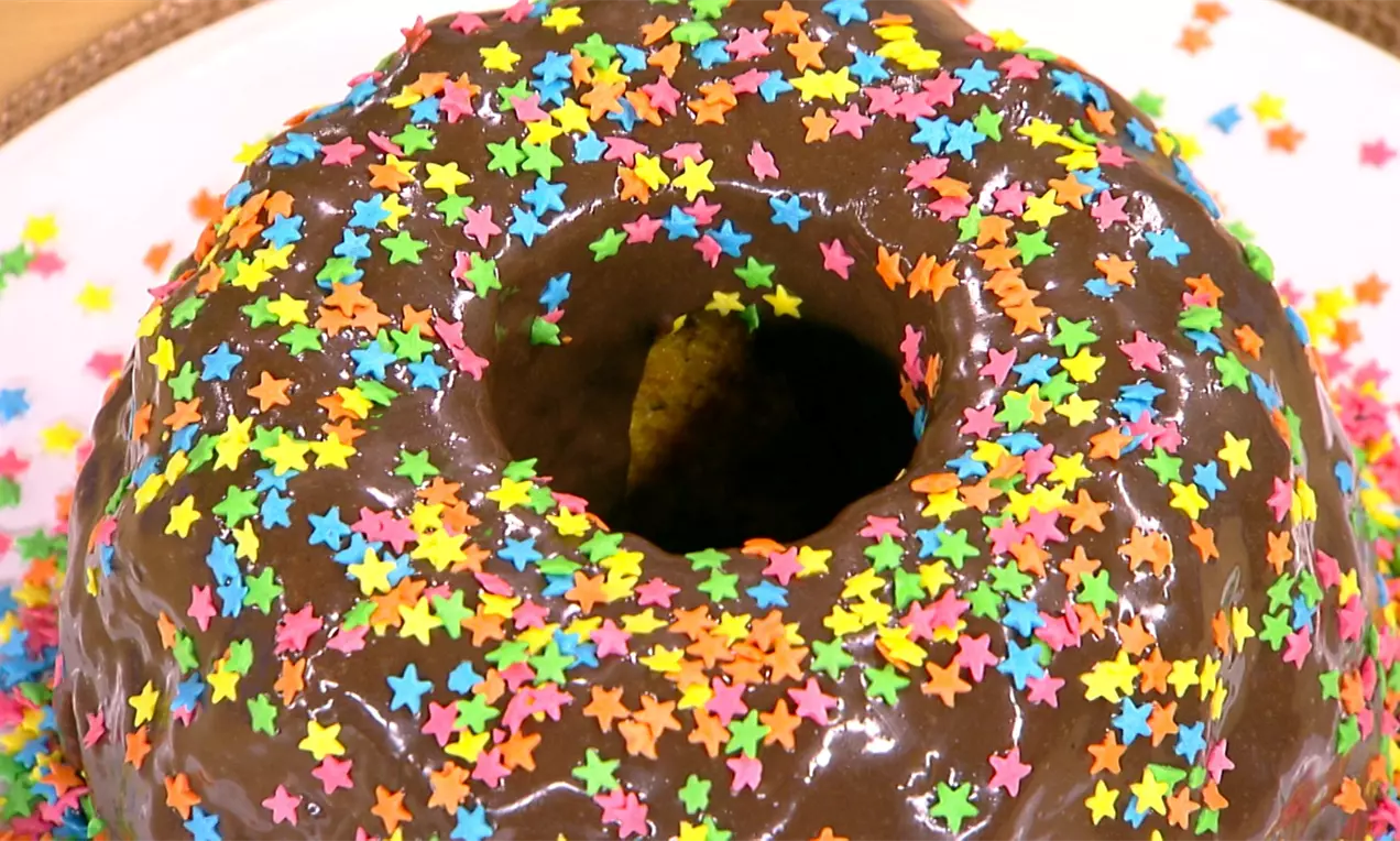Bolo circular laranja intenso com massa pontilhada de chocolate, cobertura de ganache brilhante marrom, servido em prato branco