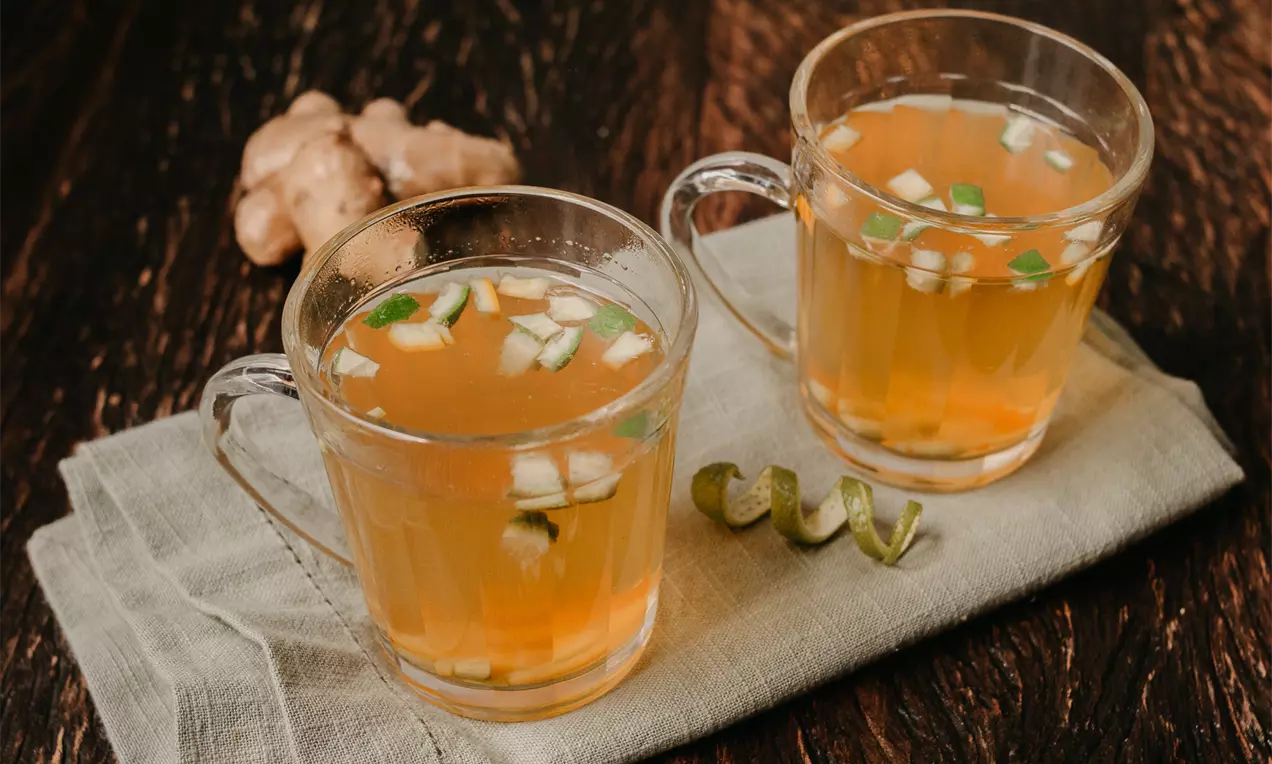 Chá alaranjado transparente com pedaços de limão, laranja e gengibre em canecas de vidro sobre guardanapo bege em mesa de madeira escura
