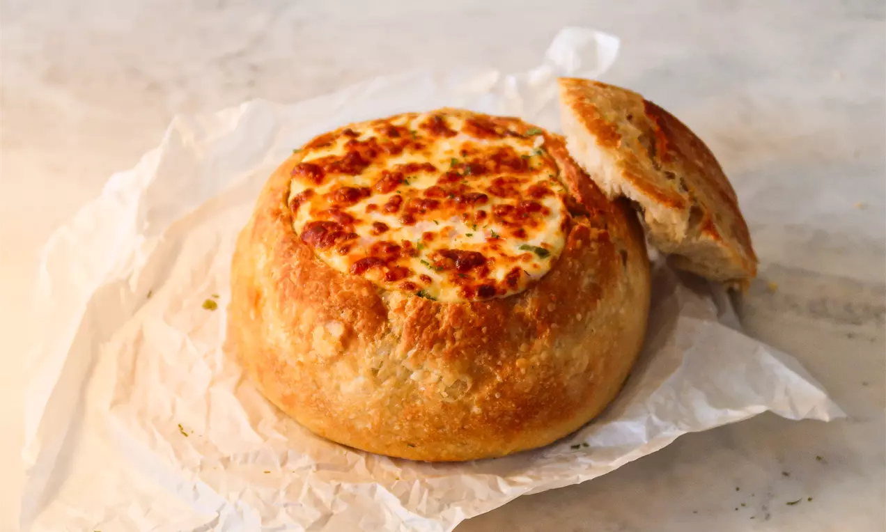 Pão redondo dourado com casca rústica e interior recheado com queijo gratinado, acompanhado de tampa cortada sobre papel branco amassado