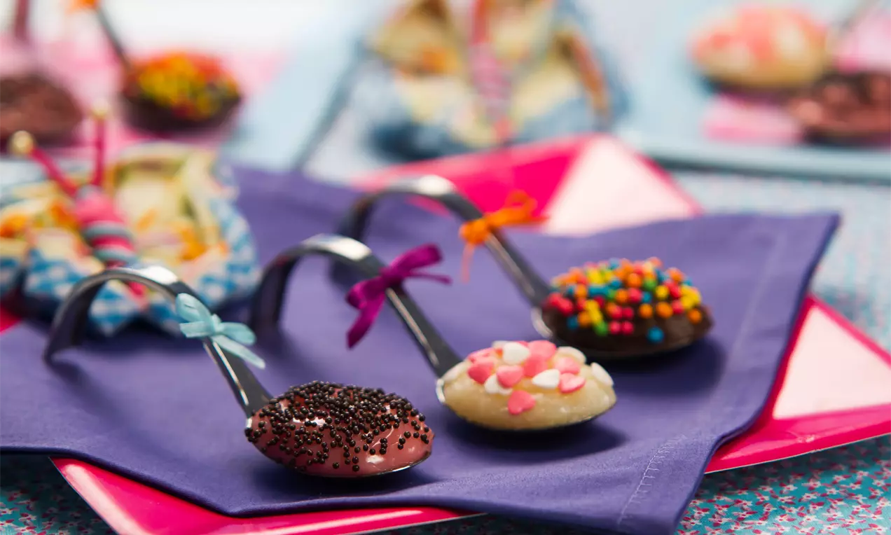Colheres de brigadeiro decoradas com miçangas coloridas sobre toalhas lilás e rosa, em um cenário vibrante e detalhado