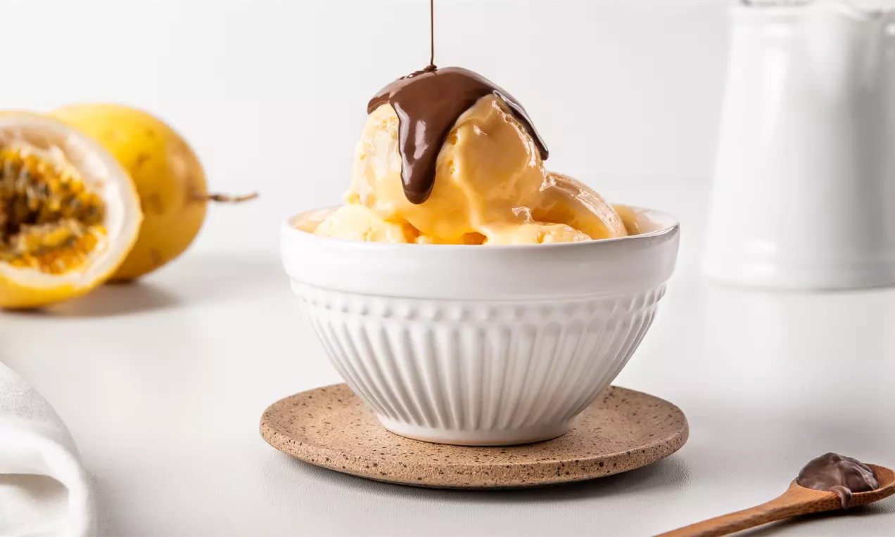 Sorvete amarelo claro de maracujá com textura suave em uma tigela branca, coberto por calda de chocolate escorrendo brilhante