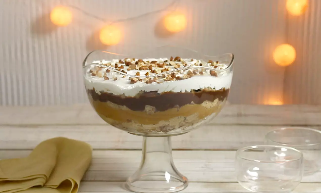 Camadas de creme dourado, chocolate brilhante e chantilly branco com cobertura de nozes picadas, servido em taça de vidro sobre madeira clara