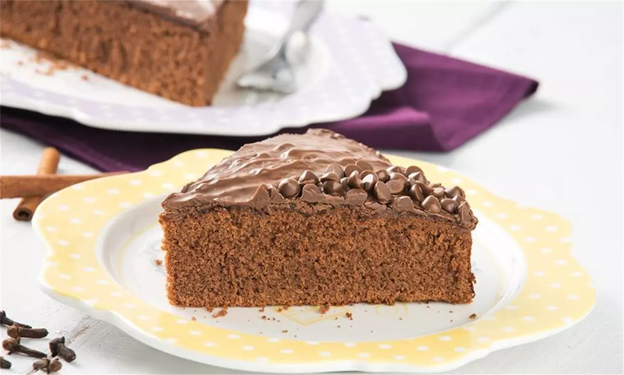 Fatia triangular de bolo pão de mel castanho, macio, com cobertura cremosa de chocolate e gotas distribuídas sobre prato amarelo com bordas brancas