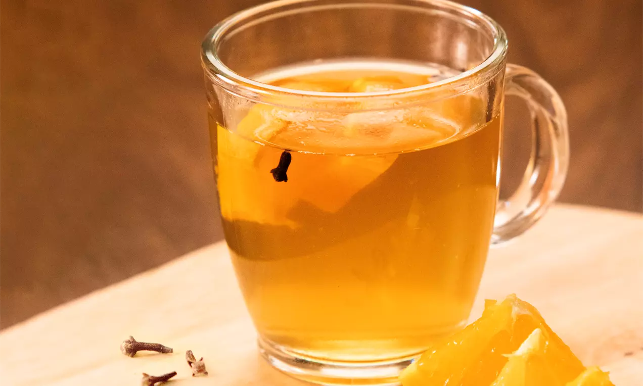 Quentão dourado translúcido em caneca de vidro, com fatias de laranja e cravos, sobre tábua de madeira com pedaços de laranja ao lado