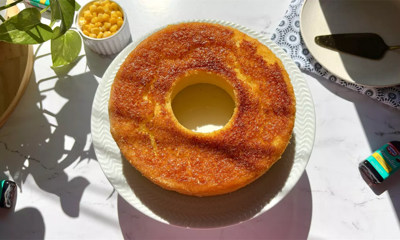 Bolo amarelo em forma de anel com crosta dourada reluzente, servido em prato branco, acompanhado por tigela de grãos de milho