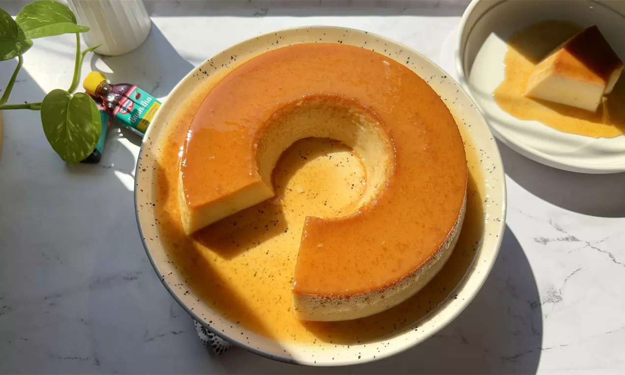 Pudim dourado em forma de anel com textura lisa e brilhante, coberto por calda caramelada, servido em prato fundo claro