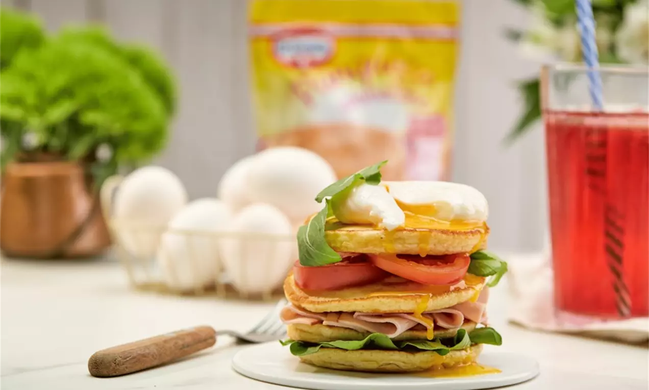 Camadas de panquecas douradas com presunto rosado, rodelas de tomate vermelho, rúcula verde e ovo pochê com gema escorrendo em prato branco