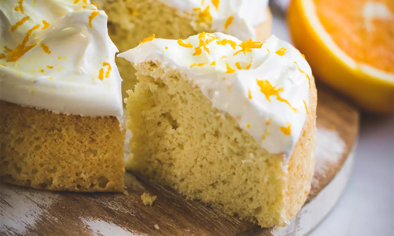 Bolo arredondado de textura macia e leve coberto com creme branco e raspas de laranja, disposto em uma tábua rústica com uma metade de laranja ao fundo