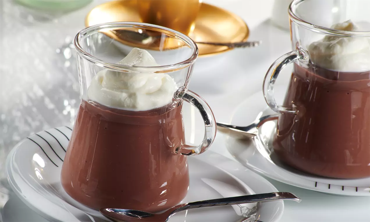 Pudim de chocolate marrom brilhante servido em xícara transparente com camada suave de chantilly branco no topo sobre prato com colher prateada