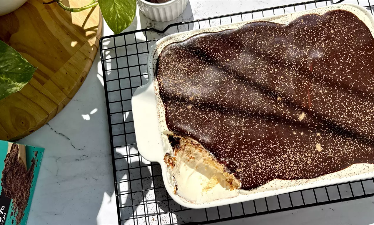 Pavê em travessa branca com camadas cremosas visíveis sob cobertura lisa de chocolate polvilhada com cacau, sobre grade de resfriamento