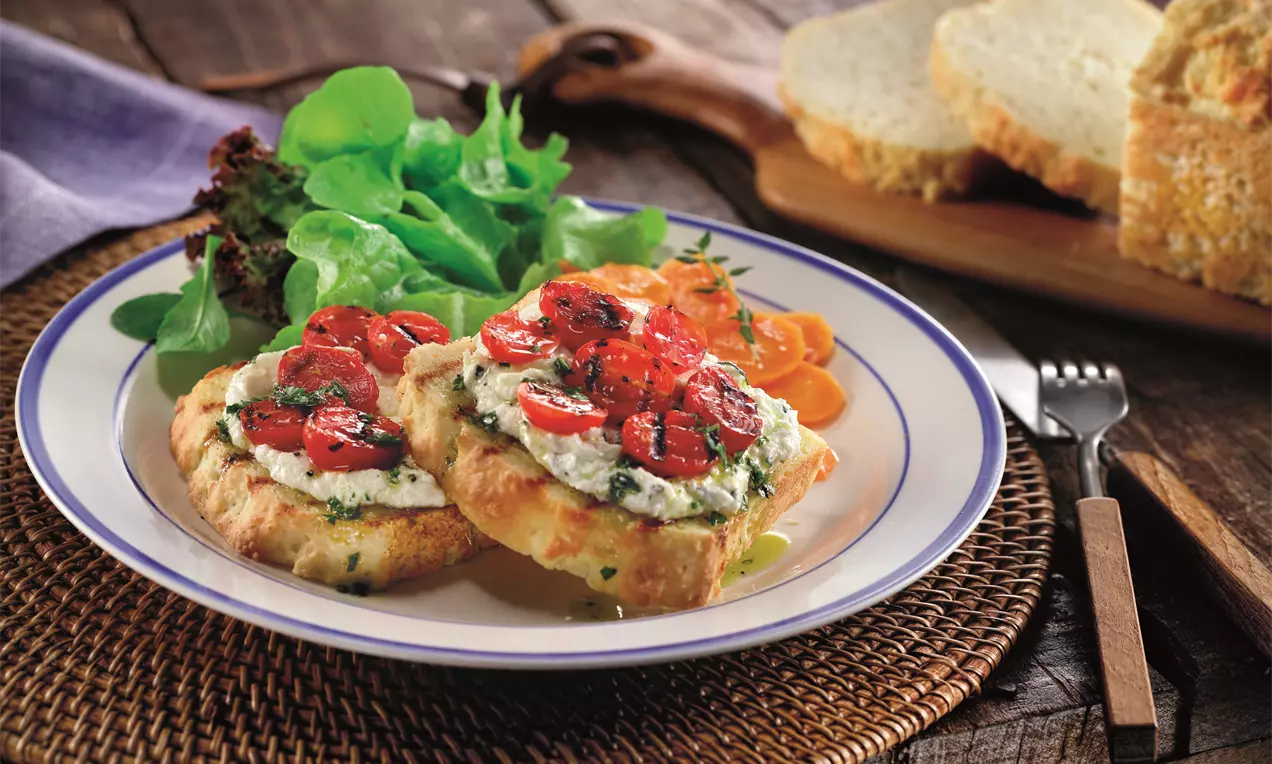 Tartine com base dourada e textura levemente crocante, coroada com creme claro e tomates assados brilhantes sobre prato cercado de folhas verdes e vegetais