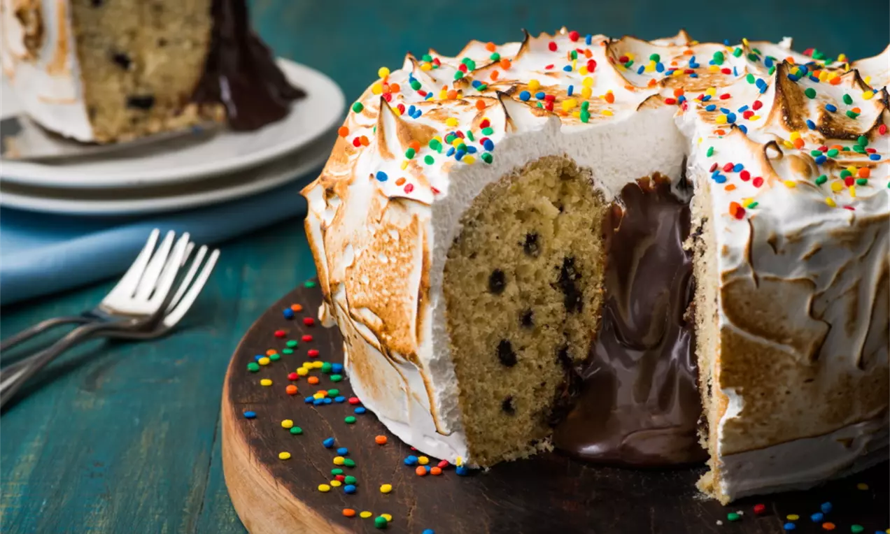 Bolo dourado com massa leve e pontos de gotas de chocolate, coberto com merengue tostado e confeitos coloridos, recheio cremoso escorrendo sobre prato de madeira