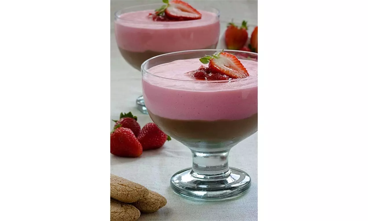 Taças de vidro com camadas de creme rosado e marrom aveludado, cobertas com morangos frescos fatiados sobre mesa com frutas e biscoitos
