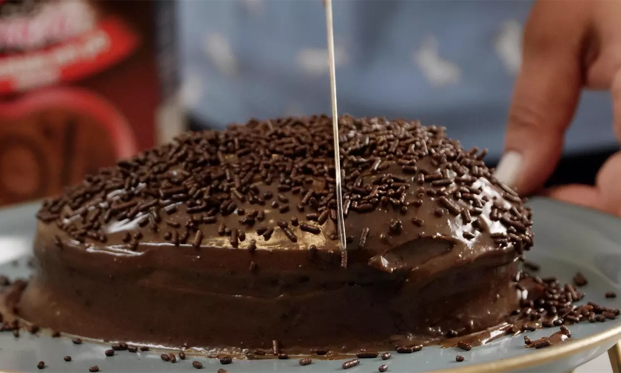 Bolo pascal coberto de ganache marrom brilhante com granulado de chocolate espalhado, servido em prato azul enquanto é cortado com faca