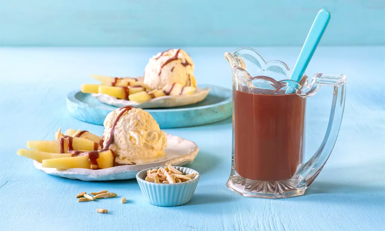Calda espessa de chocolate marrom escuro em uma jarra de vidro transparente com colher azul, ao lado de sorvetes decorados com calda e fatias de manga
