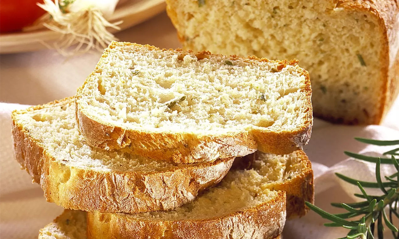 Pão italiano dourado e rústico, com crosta levemente enfarinhada e textura macia, fatiado em camadas sobre pano branco ao lado de alecrim e tomates