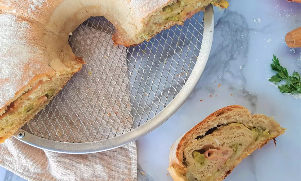 Pão dourado em forma de anel, polvilhado com farinha, recheio cremoso de abobrinha aparente, sobre uma grade metálica e toalha bege