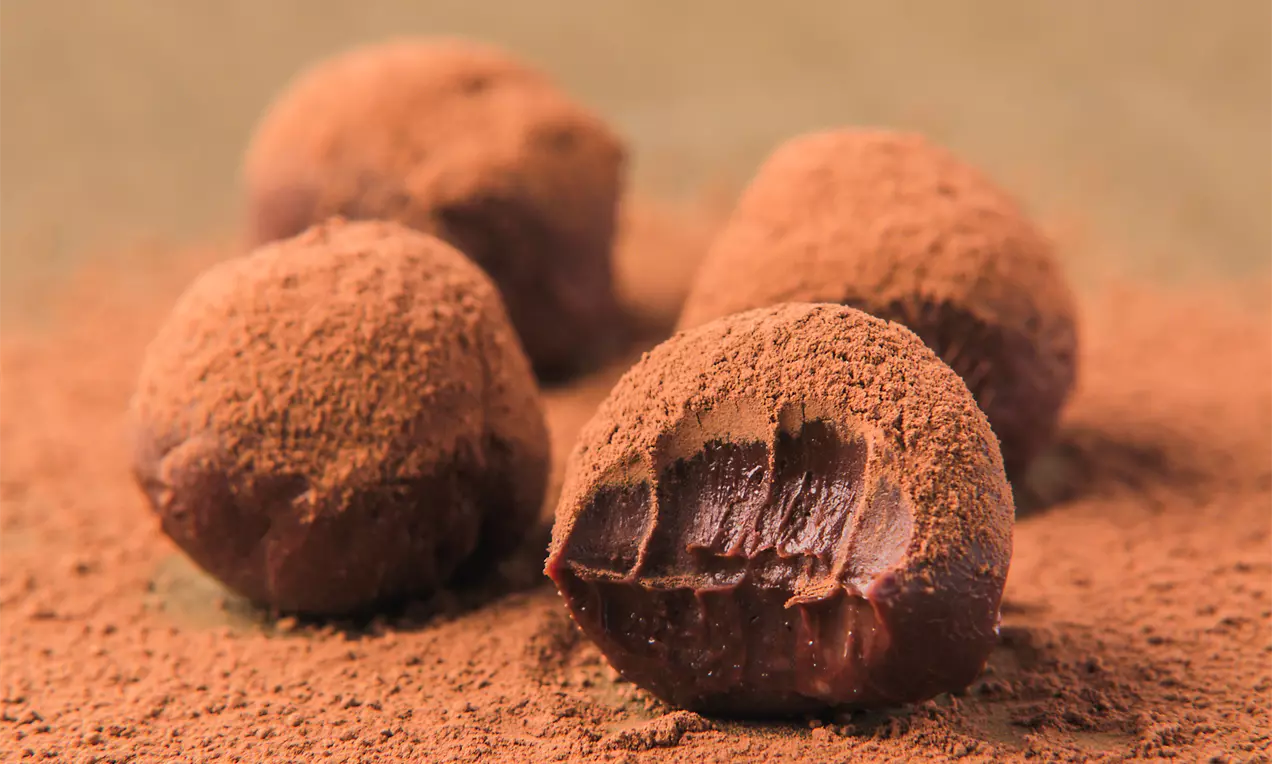 Trufas redondas de chocolate com cobertura aveludada de cacau em pó marrom, com interior macio e mordido, dispostas sobre uma camada de cacau