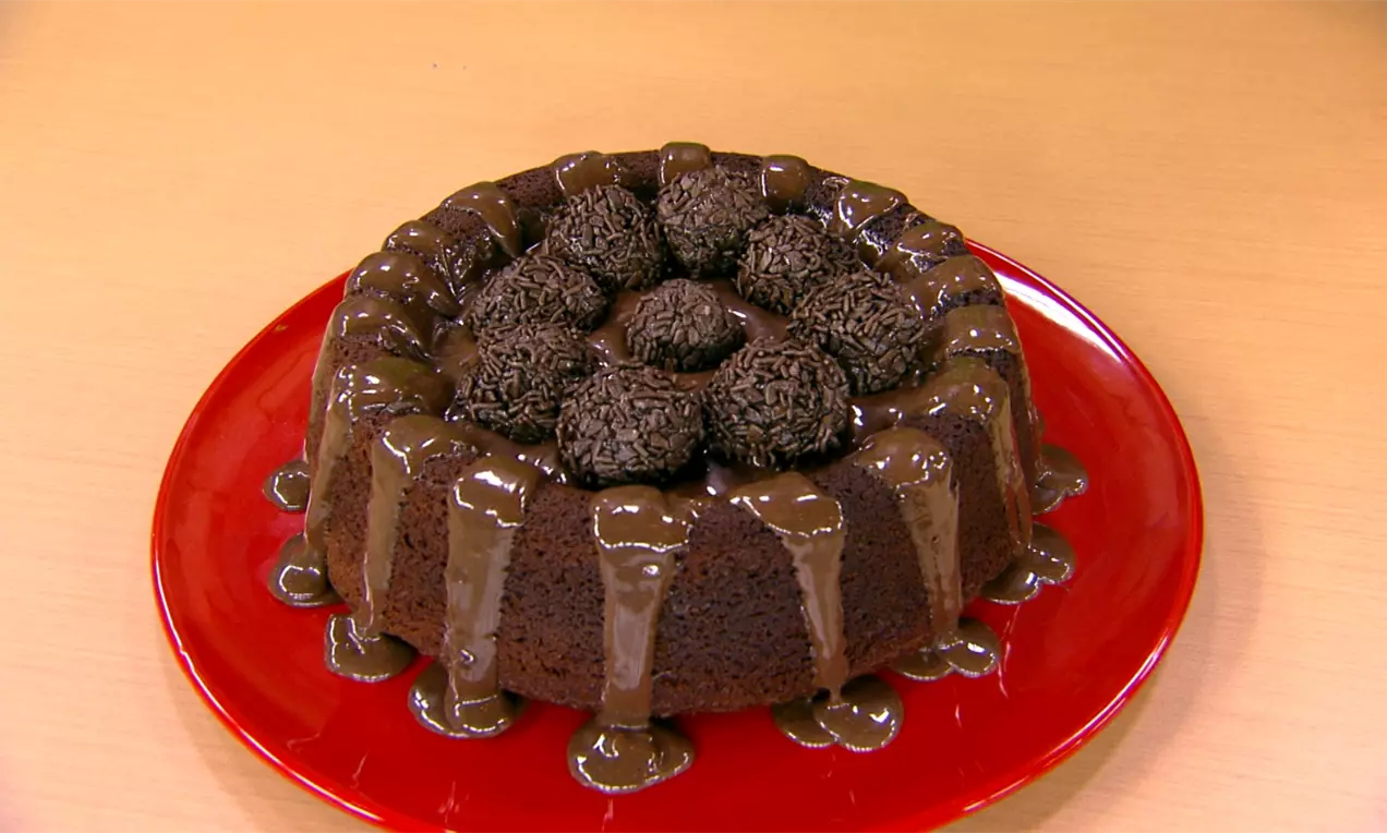 Bolo arredondado de chocolate escuro com cobertura cremosa escorrendo e decorado com brigadeiros e granulado sobre prato branco