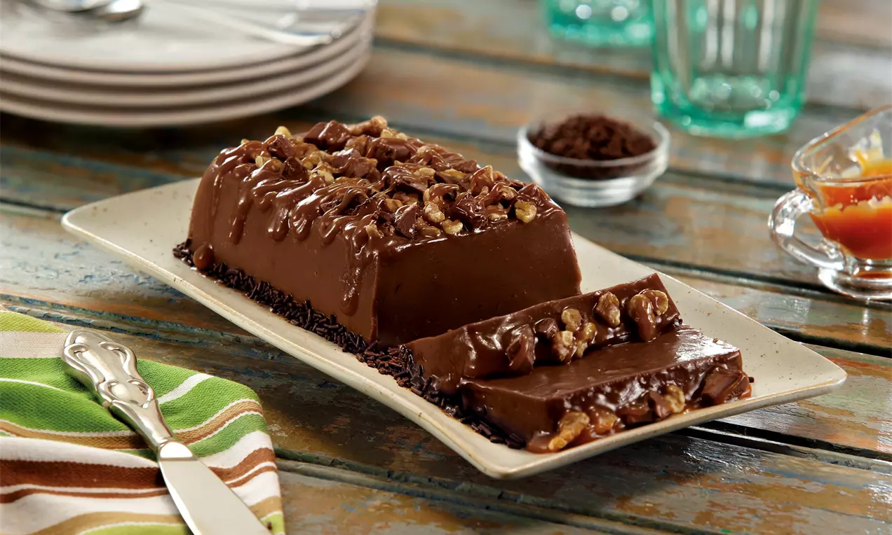 Brigadeirão retangular de chocolate brilhante com cobertura de nozes caramelizadas e borda de granulado em prato claro sobre mesa rústica