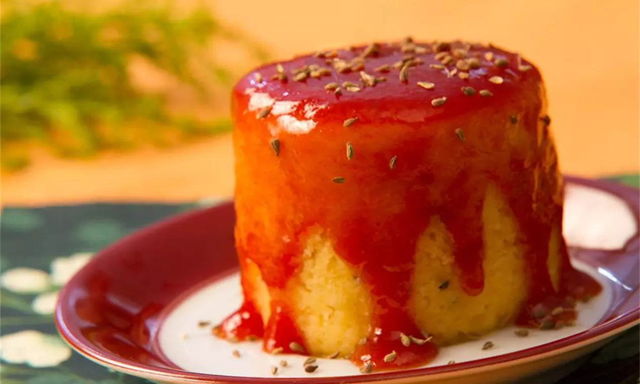 Bolo cilíndrico amarelo de textura macia coberto por geleia escarlate que escorre pelas laterais com sementes dispostas no topo em prato vermelho