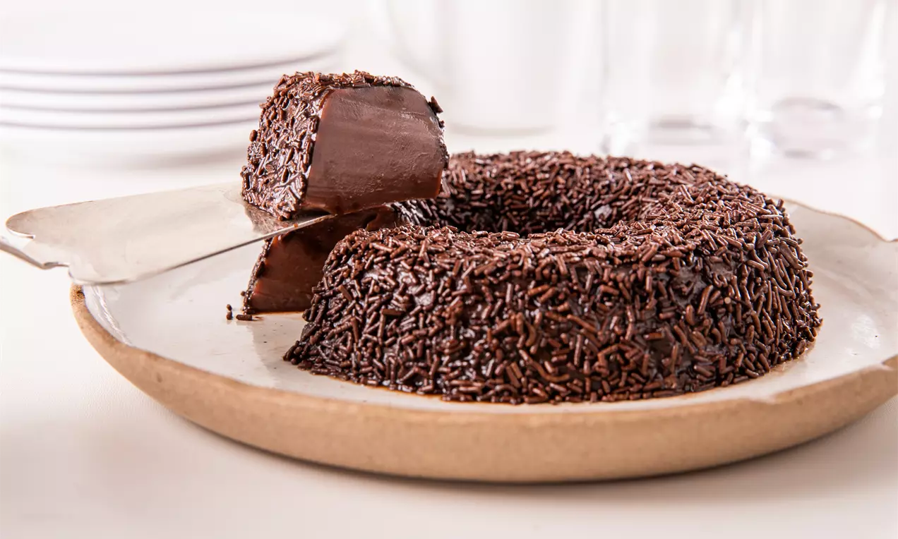 Brigadeirão cremoso com calda brilhante de chocolate, fatia removida mostrando interior chocolateado, decorado com granulado