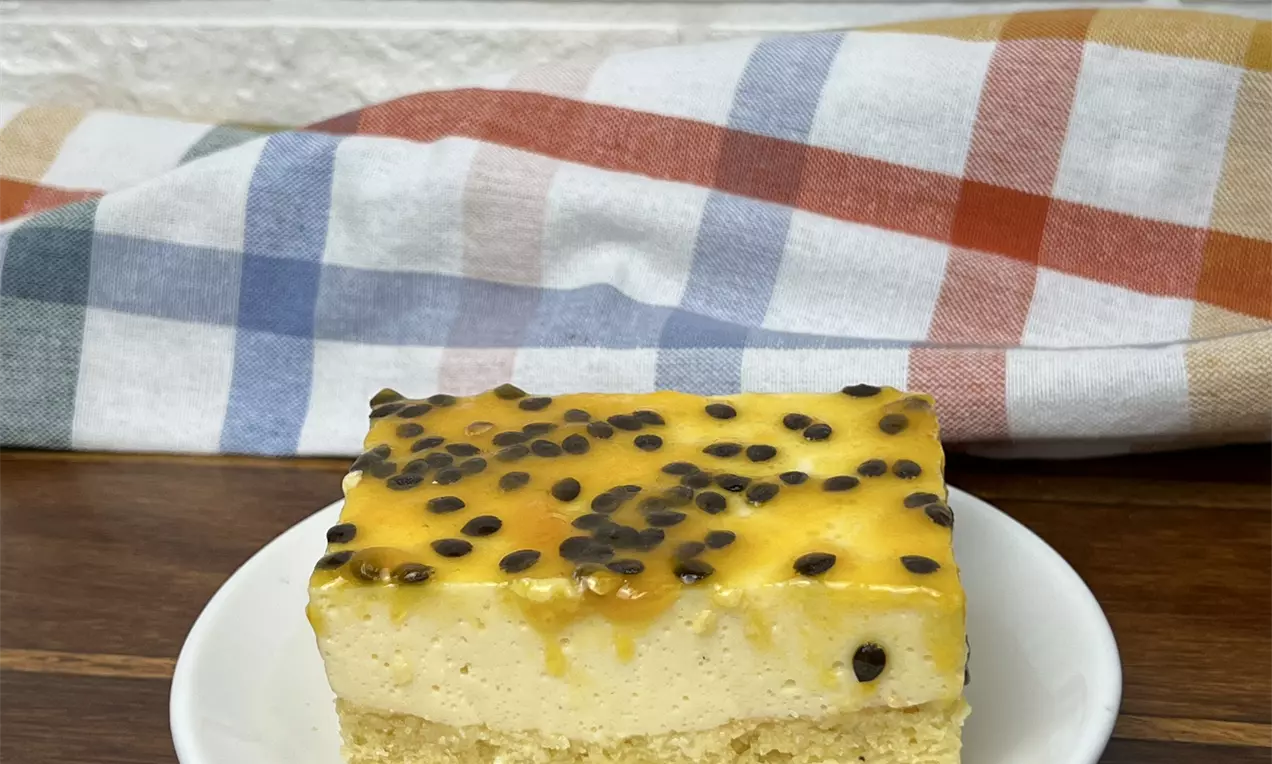 Fatia de bolo retangular com base dourada e mousse clara, coberta com geleia amarela de maracujá e sementes, servida em prato branco