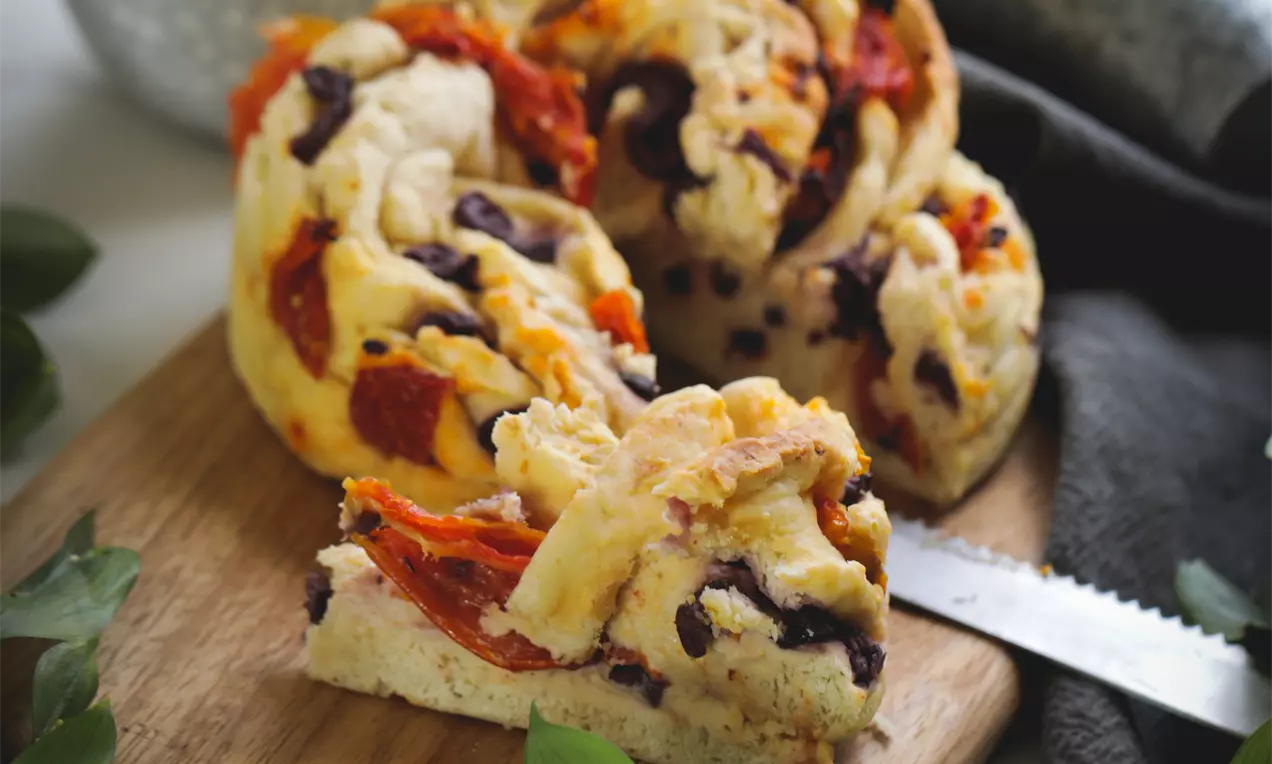 Trança dourada com textura macia e volumosa entremeada por pedaços de tomate seco e azeitonas sobre tábua de madeira cercada por folhas verdes