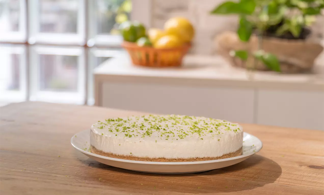 Torta sorvete de limão branca e cremosa com base fina de biscoito dourado, decorada com raspas verdes em um prato branco sobre mesa de madeira