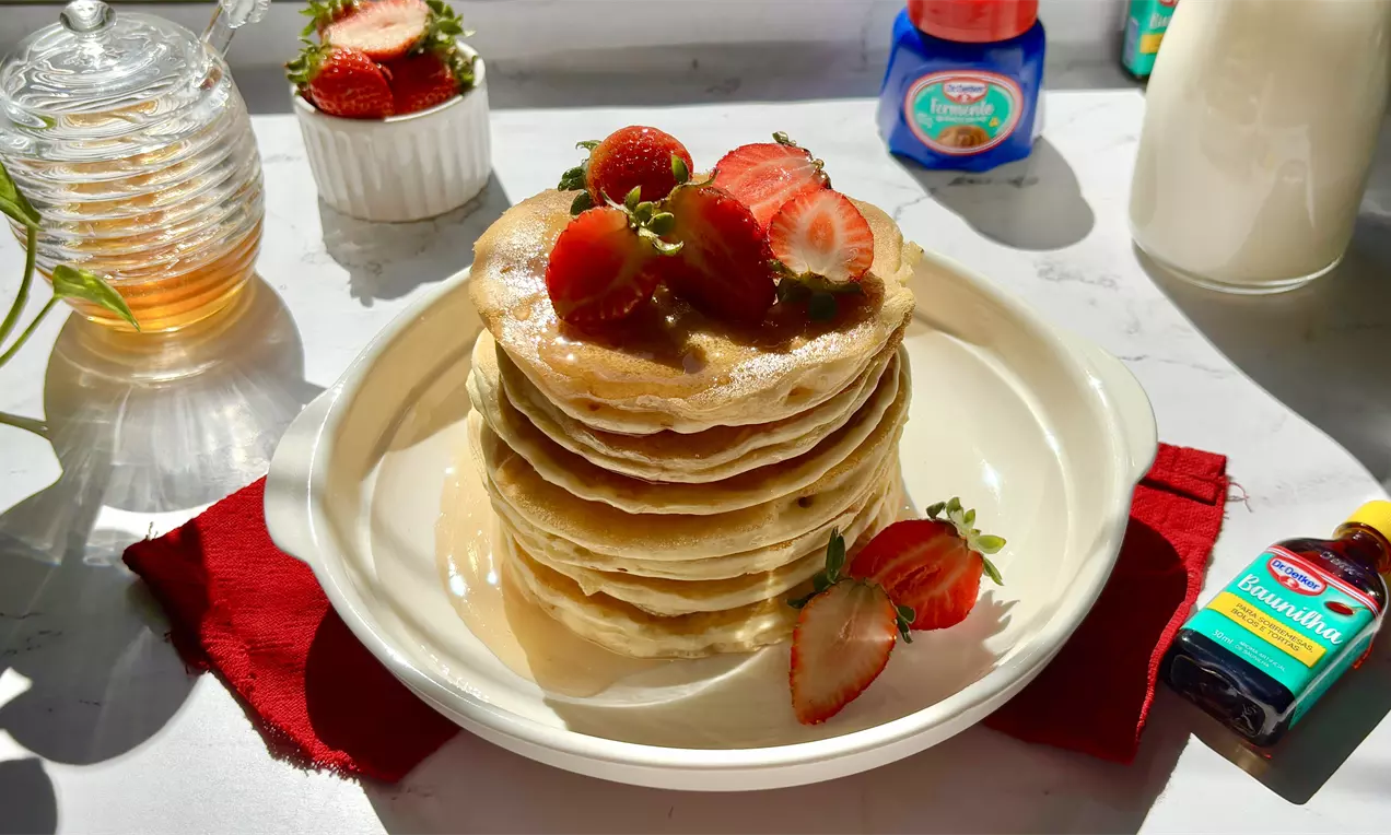 Torre de panquecas douradas com textura macia, cobertas por calda brilhante e pedaços de morango vermelho em prato branco sobre guardanapo vermelho