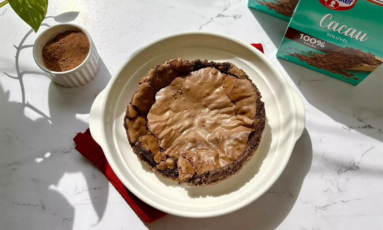 Brownie sem açúcar com superfície dourada e enrugada, textura macia ao centro e bordas crocantes, servido em prato branco sobre toalha vermelha