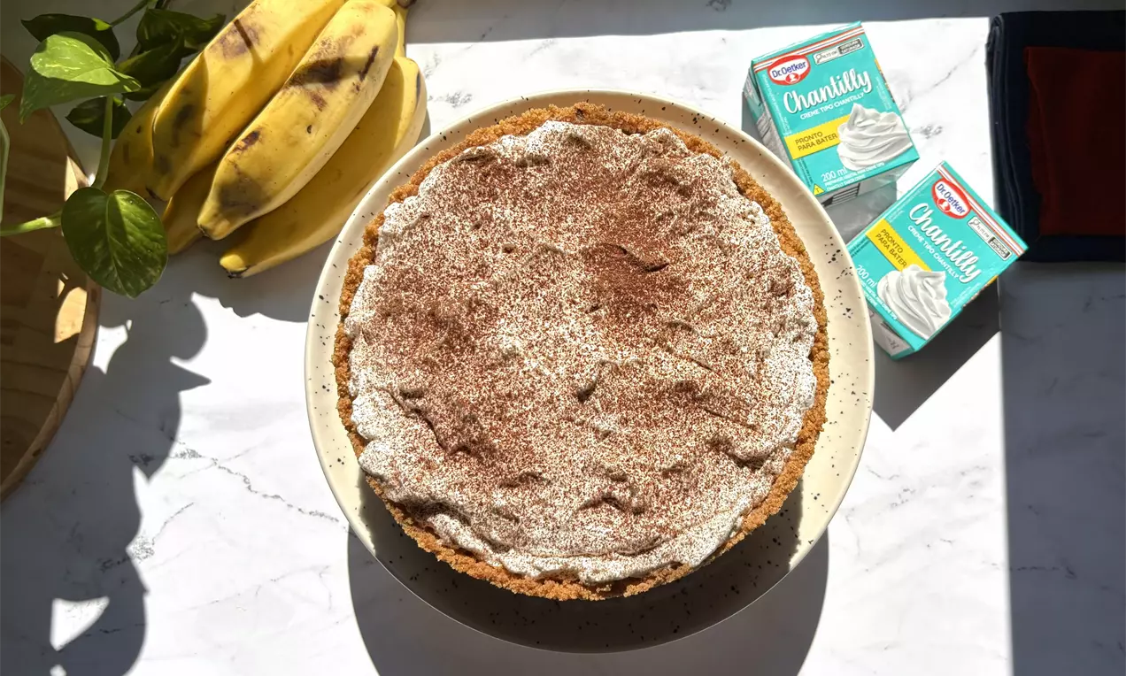 Torta com base marrom de biscoito crocante, cobertura cremosa clara polvilhada com cacau em pó, ao lado de bananas e creme chantilly