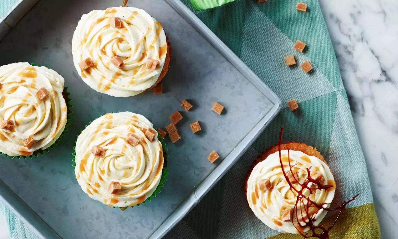 Muffins dourados com cobertura espiral branca, fios de caramelo e cubos de doce em uma bandeja cinza sobre tecido geométrico