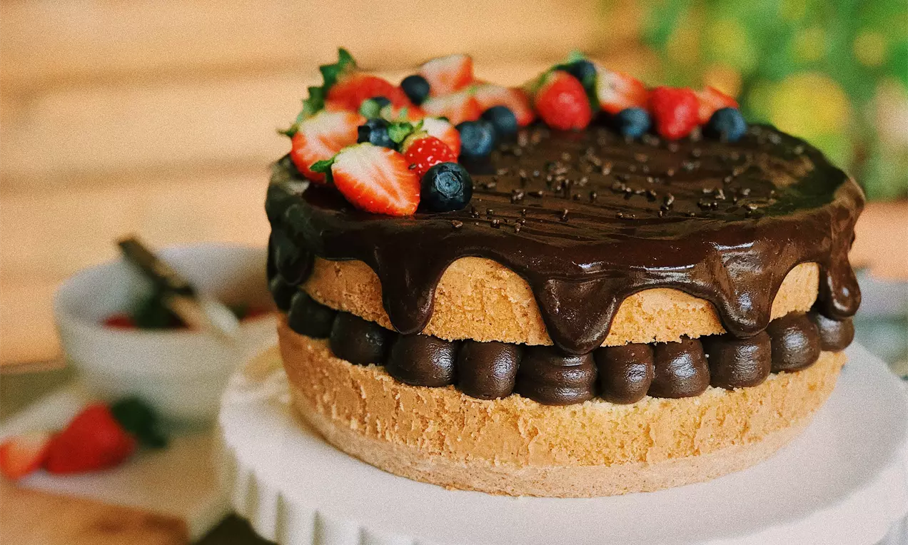 Bolo trufado coberto por ganache de chocolate escuro, decorado com morangos e mirtilos frescos sobre prato branco redondo