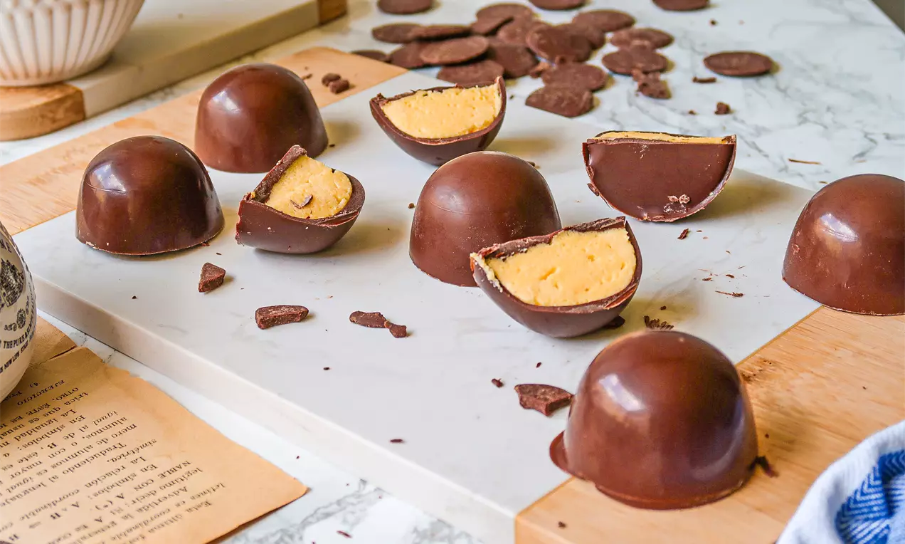 Bombom em formato semiesférico com casca de chocolate brilhante e recheio cremoso amarelo intenso, disposto sobre tábuas e mármore branco