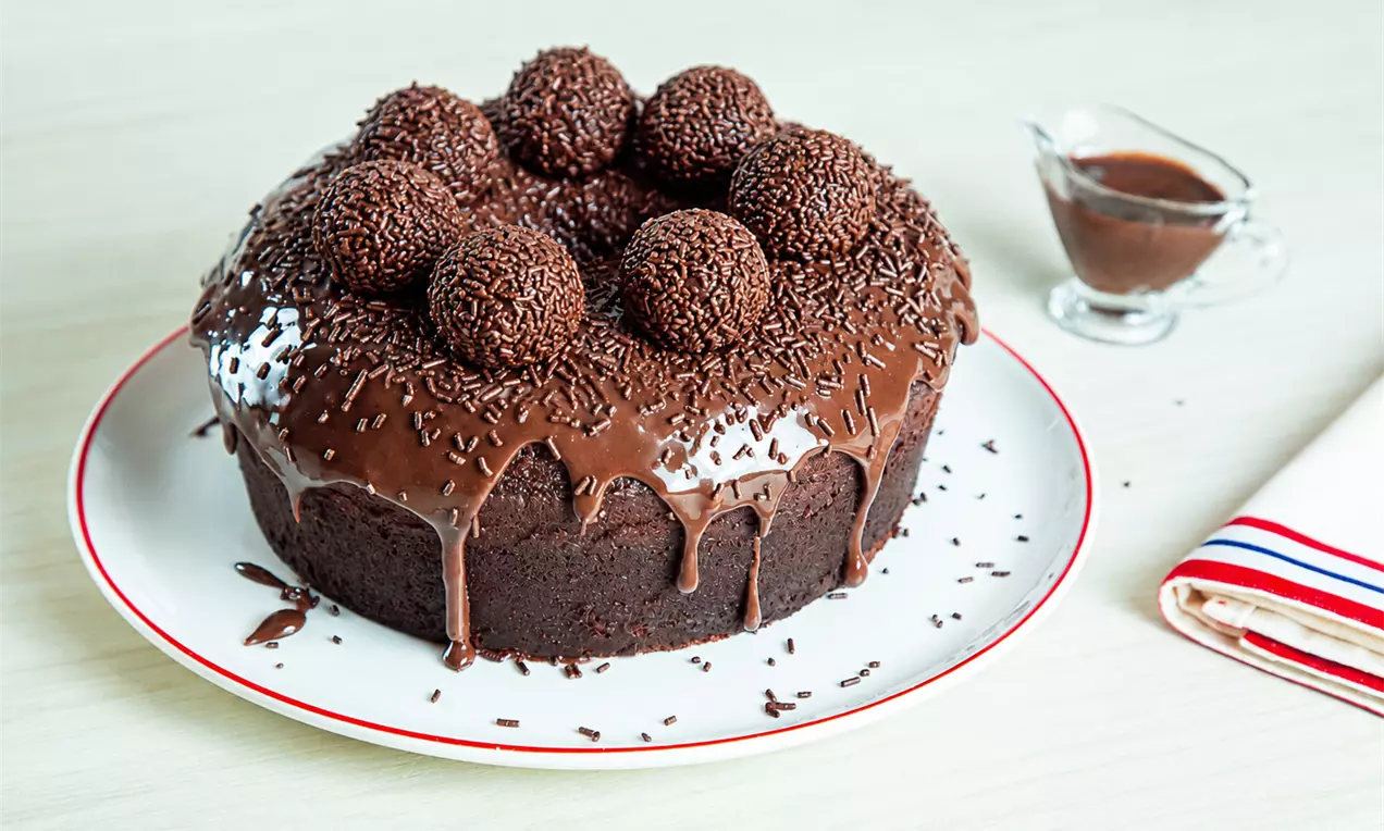 Bolo arredondado de chocolate escuro com cobertura cremosa escorrendo e decorado com brigadeiros e granulado sobre prato branco