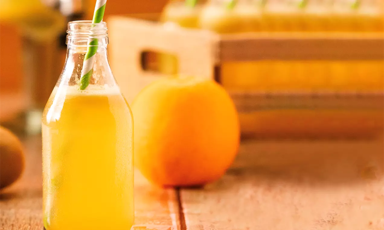 Garrafa de vidro transparente com chá amarelo dourado e canudo verde listrado sobre mesa rústica ao lado de uma laranja inteira