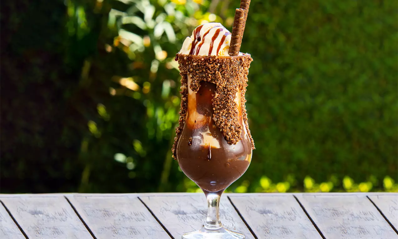 Taça com borda coberta de chocolate e granulados, recheada com creme escuro, pedaços de doce, bola de sorvete e canudo de chocolate