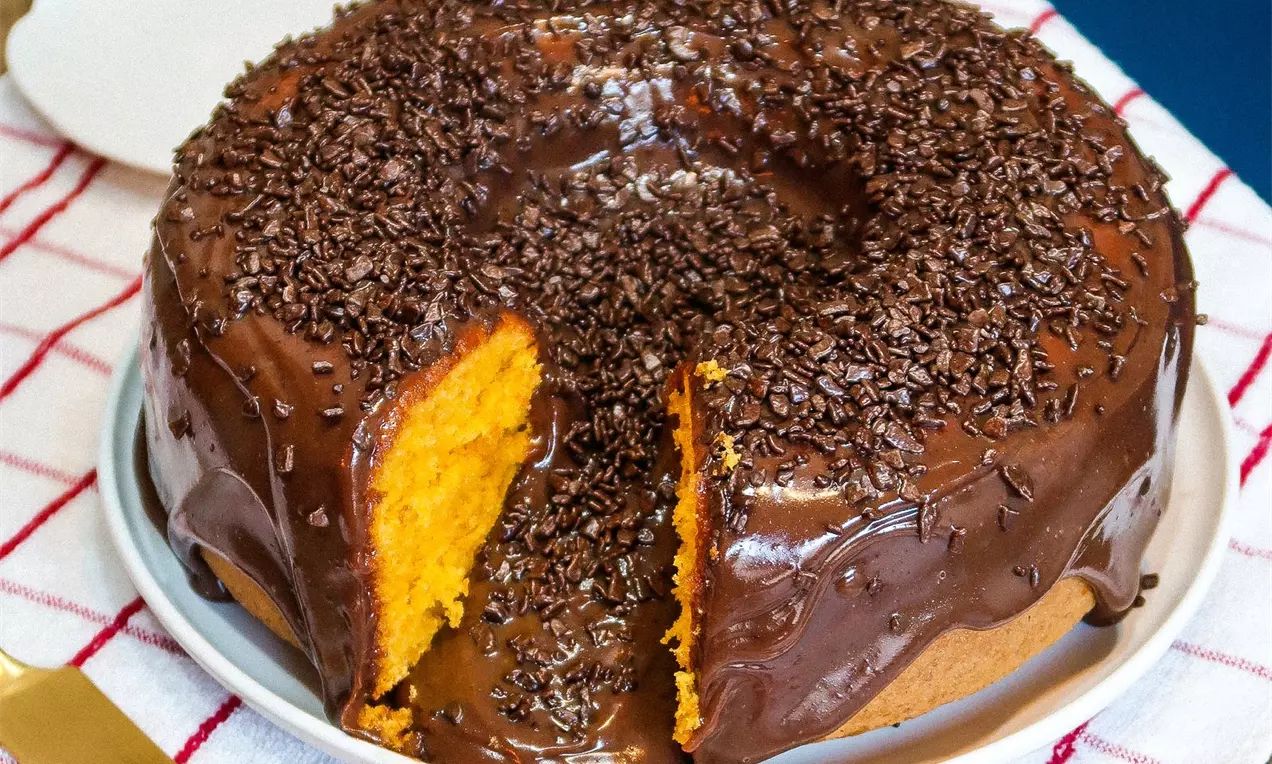 Bolo redondo de cenoura alaranjado com cobertura espessa de chocolate brilhante e raspas finas sobre um prato branco com pano listrado vermelho