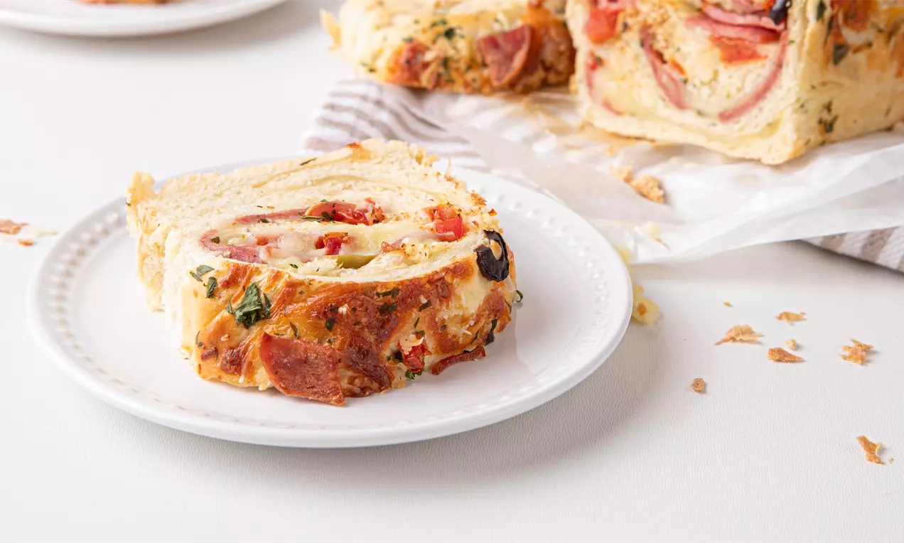 Pão macio e dourado com recheio de calabresa, queijo derretido e ervas visíveis em fatia redonda sobre prato branco próximo a pedaços soltos