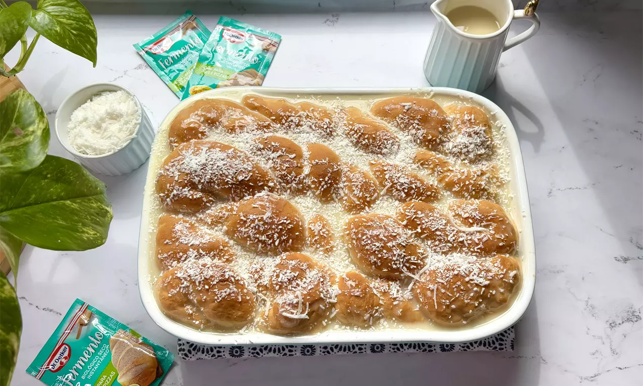 Pães dourados e macios dispostos em uma travessa branca, cobertos com calda cremosa e salpicados com coco ralado branco