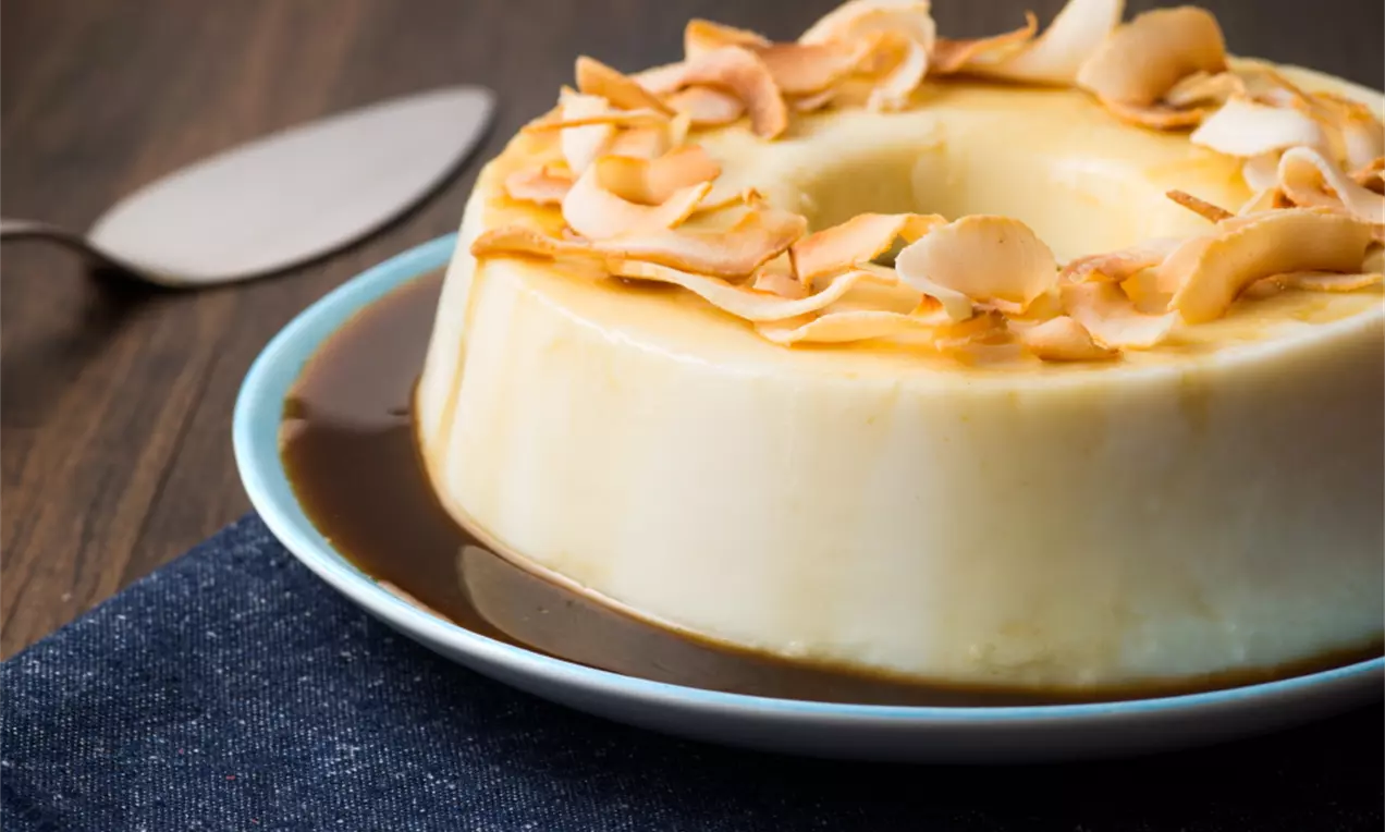 Manjar de coco branco brilhante em formato de anel, com uma calda escura de melado ao redor e lascas douradas de coco queimado no topo