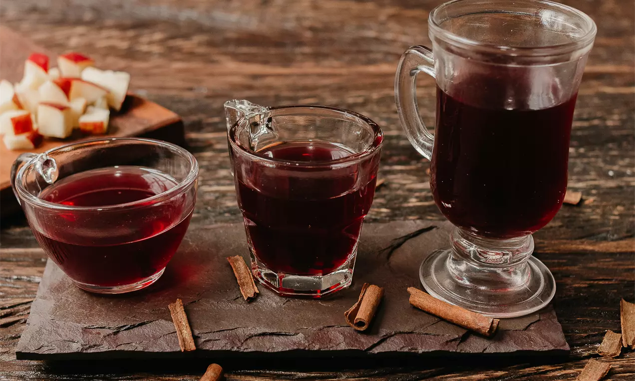 Vinho quente rubi-escuro servido em copos de vidro transparentes sobre base de ardósia com paus de canela e cubos de maçã ao fundo