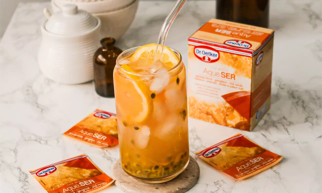 Drink dourado translúcido com gelo, fatias de laranja e sementes de maracujá em copo alto com canudo sobre base de mármore claro