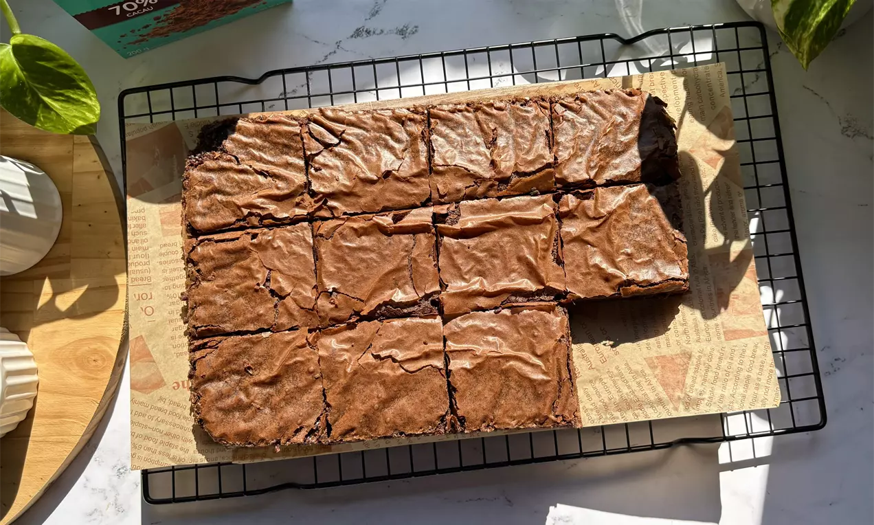 Brownie retangular com superfície marrom brilhante e rachaduras finas, cortado em pedaços iguais sobre papel manteiga e grade metálica