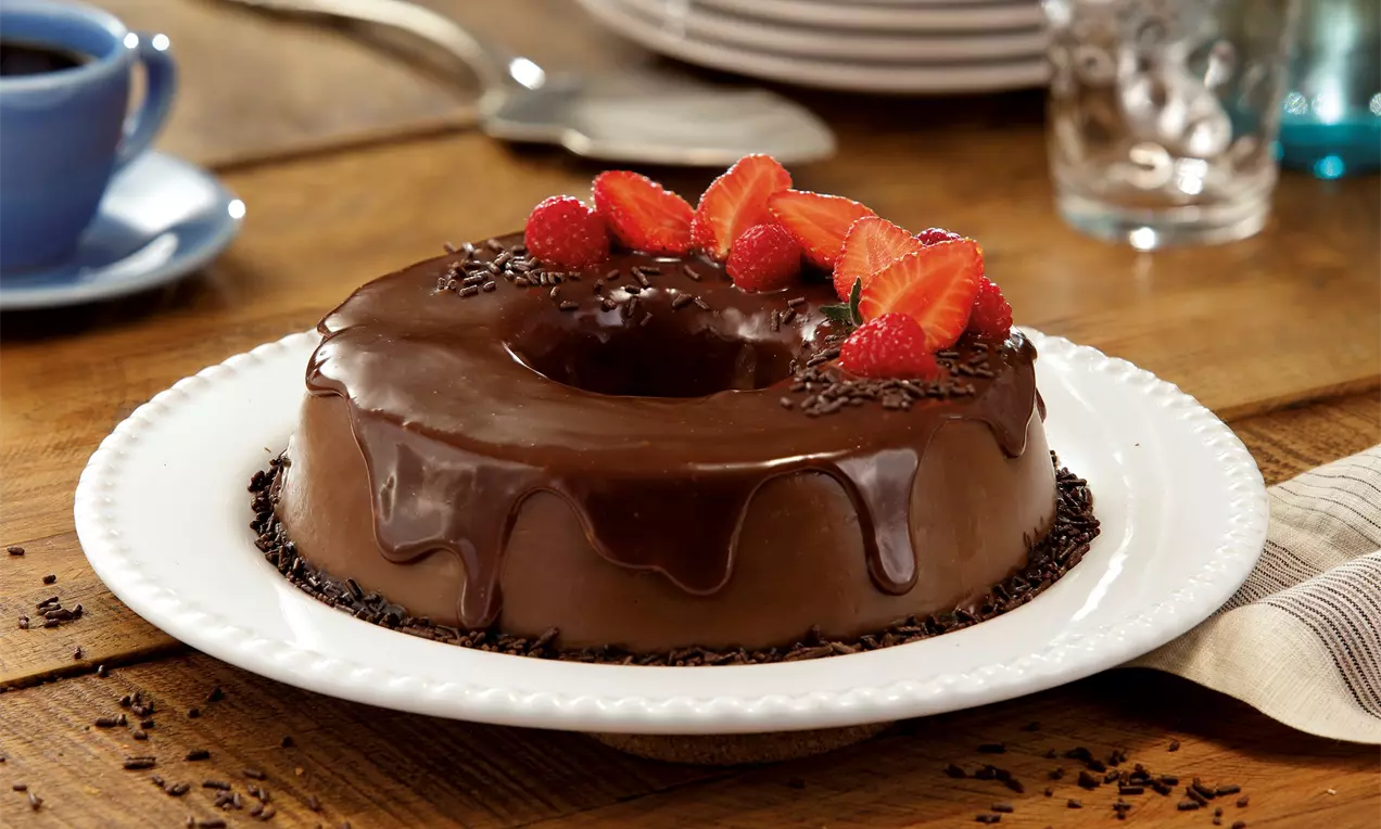Brigadeirão escuro e brilhante com ganache escorrendo, decorado com morangos vermelhos e granulado em prato branco sobre mesa de madeira