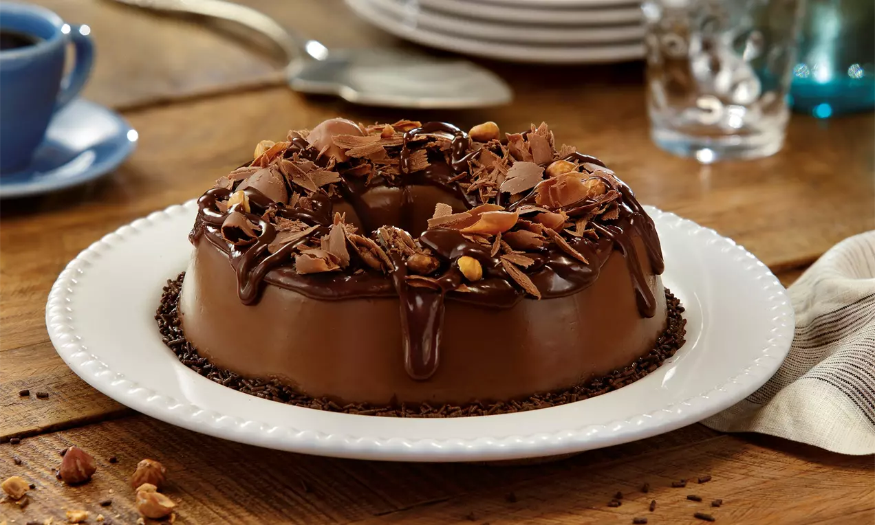 Brigadeirão brilhante de chocolate marrom decorado com lascas de chocolate, avelãs inteiras e calda escura em um prato branco redondo